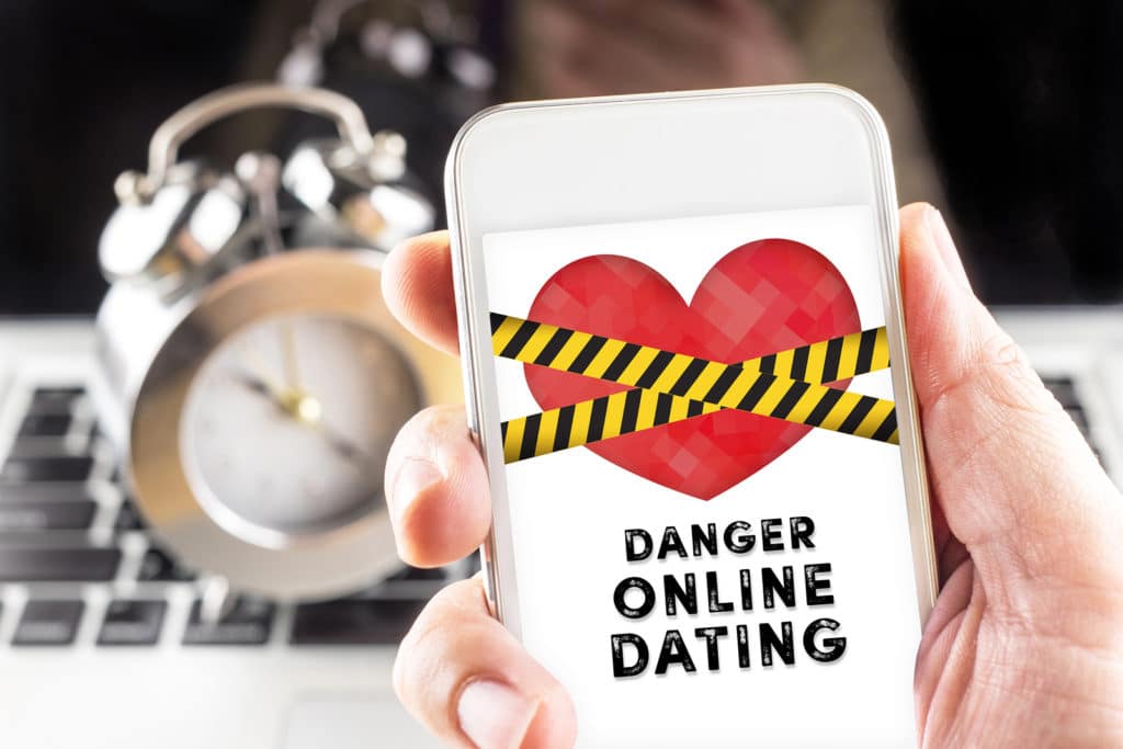 Hand holding mobile with caution tape on heart and Danger online dating on screen with clock and laptop at background Internet love concept.