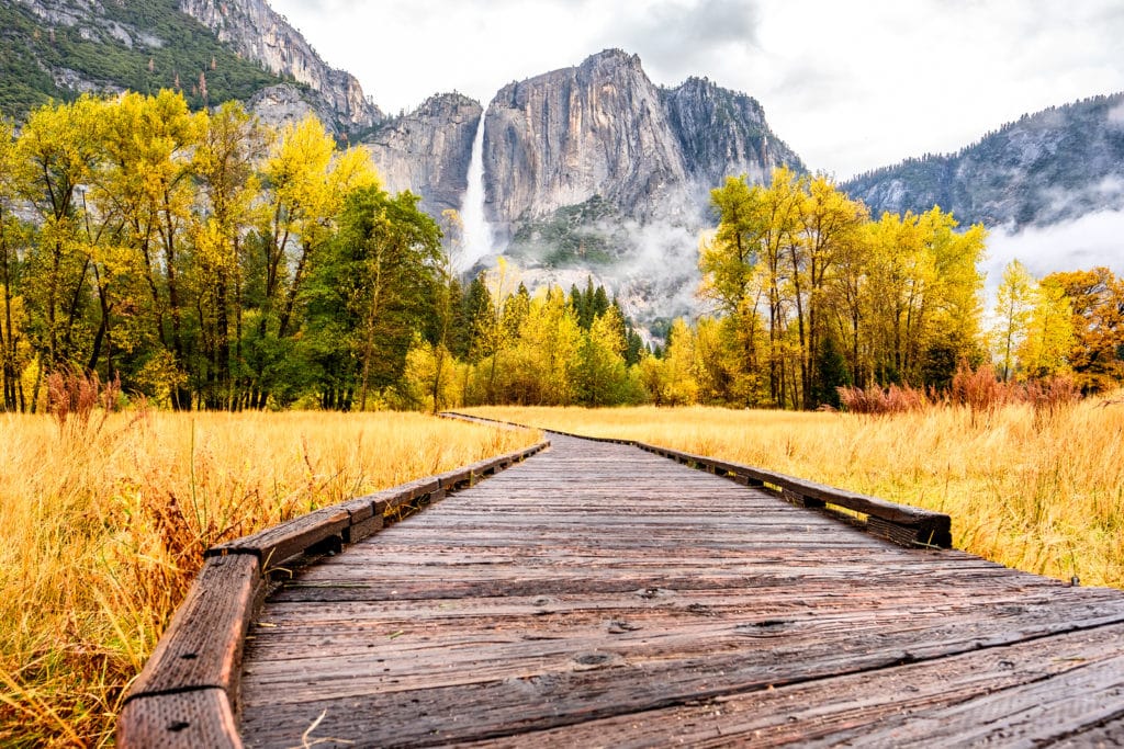 Meadow with boardwalk in Yosemite National Park Valley with Yosemite Falls at cloudy autumn morning. Low clouds lay in the valley. California, USA.