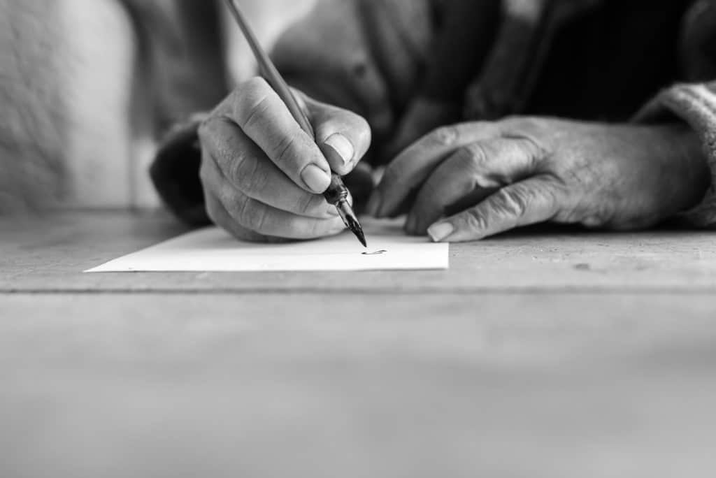 Greyscale image of an elderly man doing calligraphy writing using a nib pen and ink on a sheet of white paper in a low angle close up view of his hands with foreground copy space.